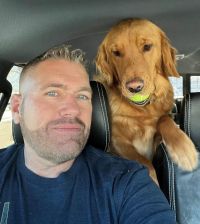 John Burfeind - this is a selfie taken by John in his truck. He has short hair, shaved at the sides, blue eyes, and pronounced stubble. He is wearing a dark blue shirt. His Golden Retriever dog is above his left shoulder, facing the camera, and has a tennis ball in its mouth.